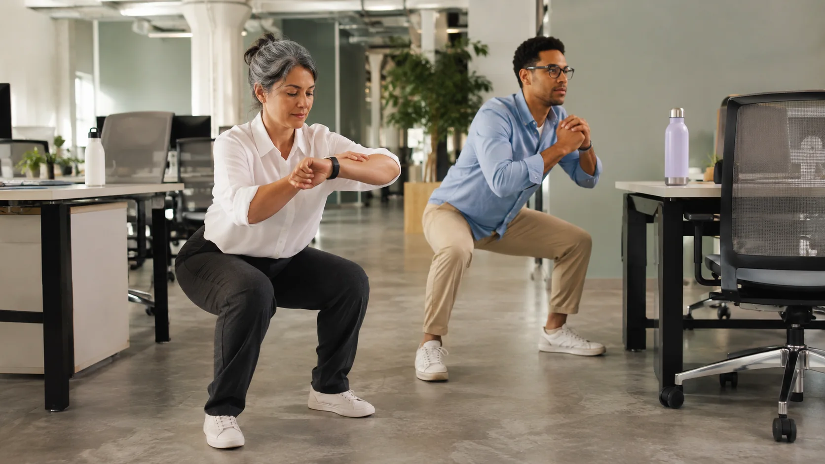 Two office workers performing brief chair squats beside their desks