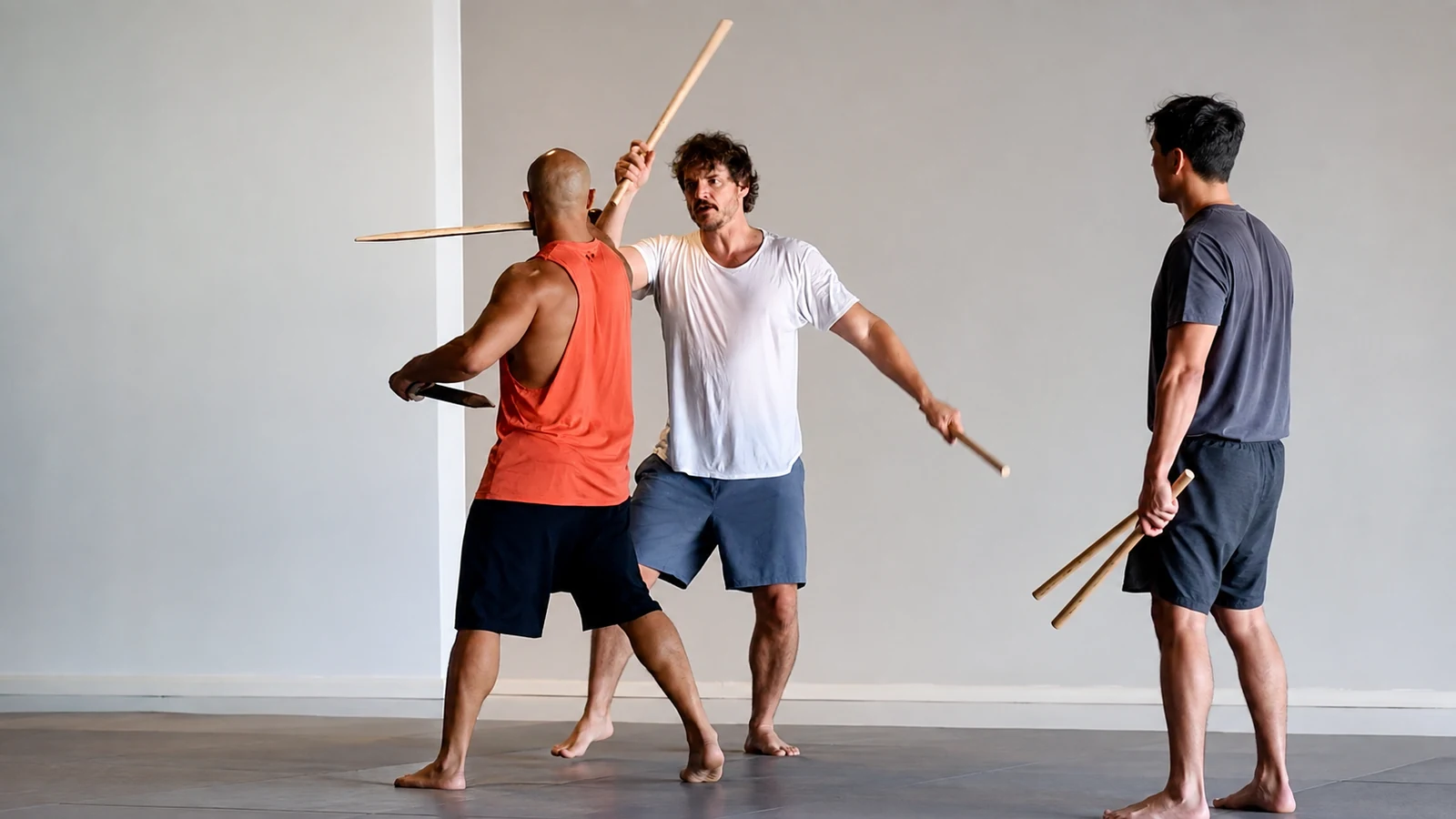 Pedro Pascal rehearsing controlled movement with training partners and padded wooden sticks in a bright studio