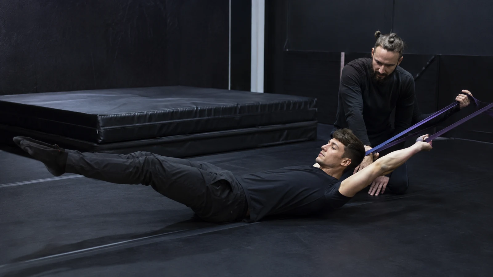 Male performer holds a hollow-body position with resistance-band shoulder work while a coach checks alignment beside crash mats.