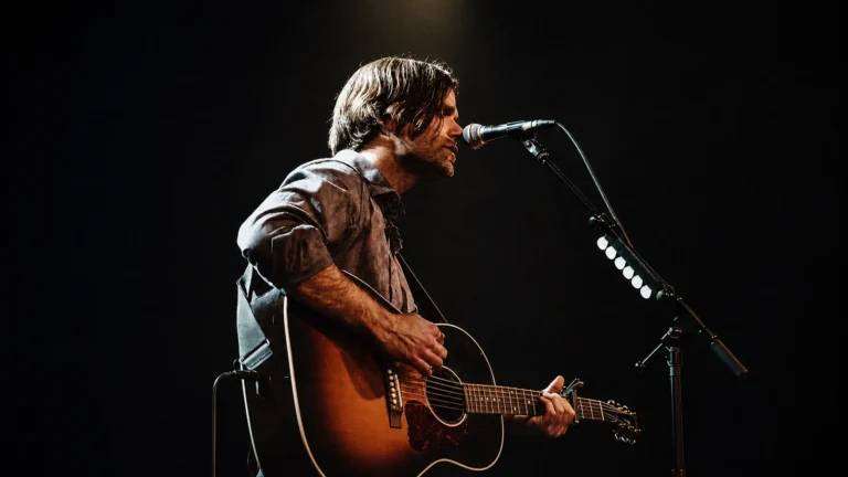 Ben Gibbard singing into a microphone while playing acoustic guitar onstage