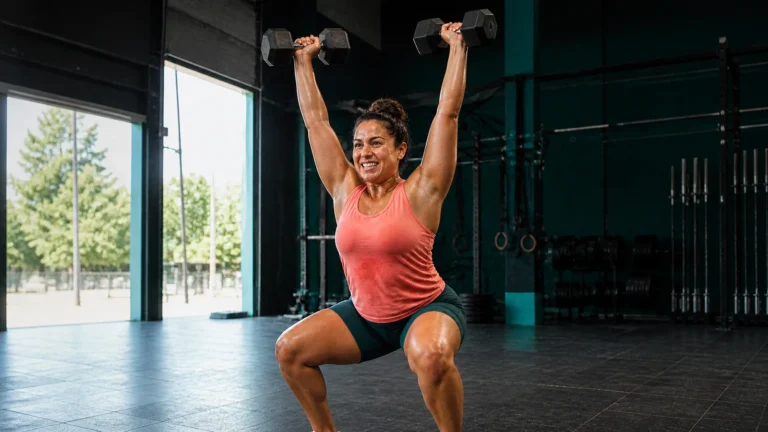 Woman in a coral top holding two dumbbells overhead in a teal gym.