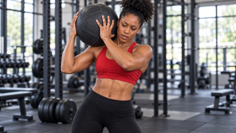 Woman in a red athletic top holding a black medicine ball beside her shoulder in a gym.