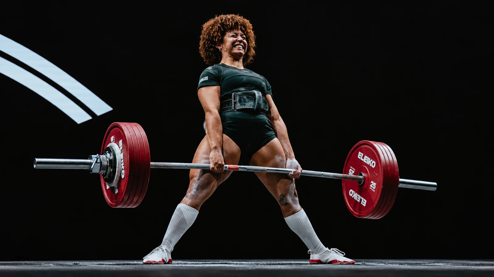 Powerlifter in a dark green singlet holding a barbell at deadlift lockout on a dark competition platform.