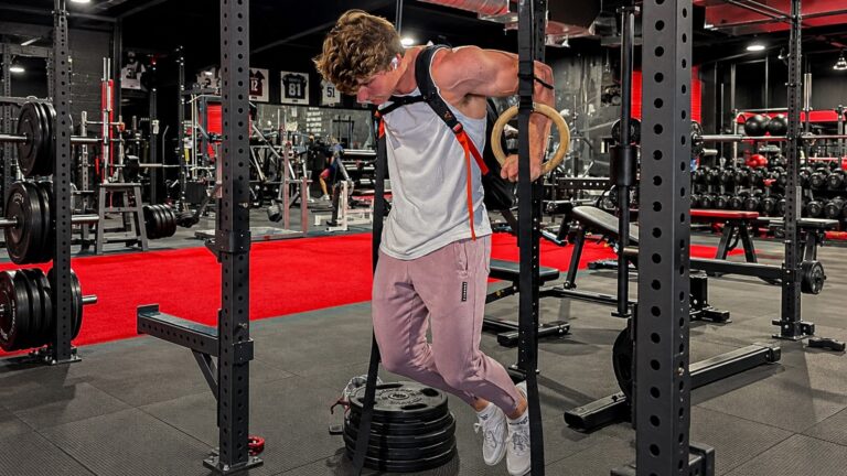 Jake Rintoul performing a weighted ring dip in a gym, wearing a backpack with gymnastic rings beside a squat rack.